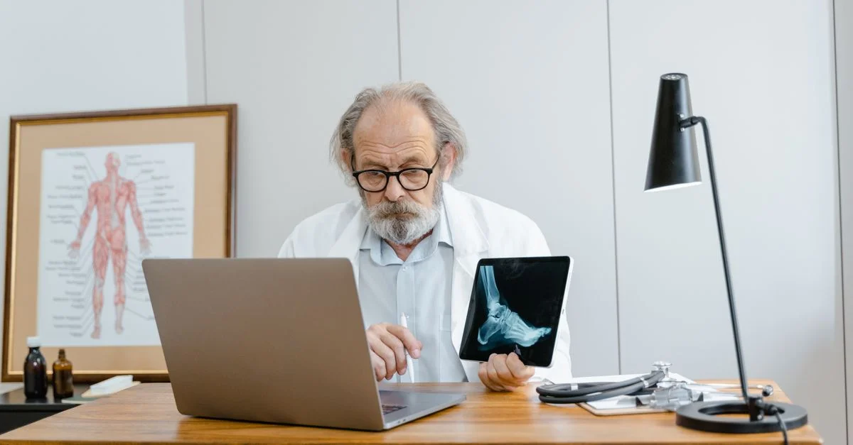 Médecin masculin âgé examine une radiographie sur une tablette tout en travaillant à son bureau avec un ordinateur portable.
